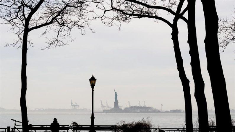 Una persona se sienta en un banco en una sección casi vacía de Battery Park con la Estatua de la Libertad al fondo en Nueva York, Nueva York, EE.UU., el 20 de marzo de 2020. EFE/EPA/JUSTIN LANE
