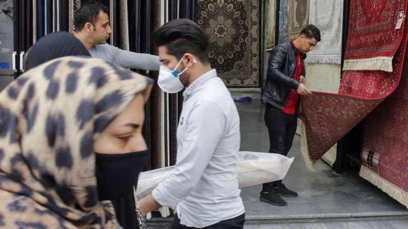 Iraníes, algunos de ellos con máscaras protectoras, caminan frente a una tienda de alfombras en las afueras del gran bazar de la capital Teherán, durante las crisis de la pandemia del virus del PCCh, el 18 de marzo de 2020. (Foto de -/AFP vía Getty Images)