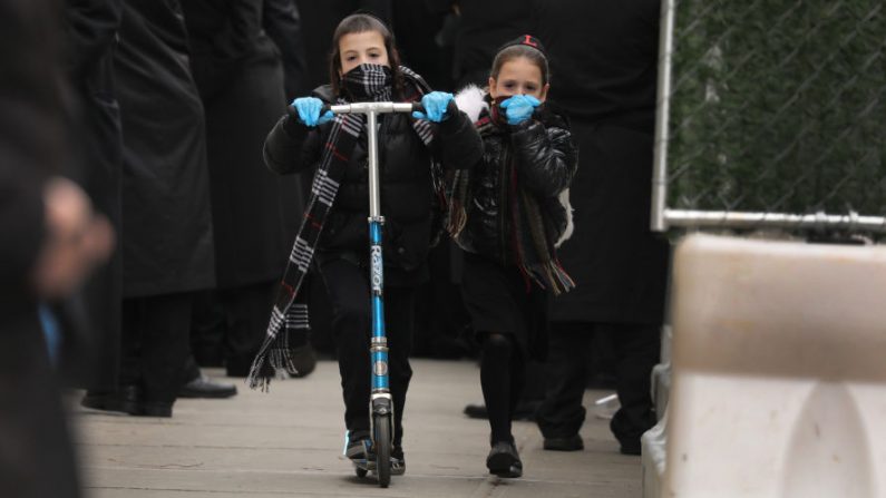 Niños viajan en scooters con máscaras faciales mientras cientos de miembros de la comunidad judía ortodoxa asisten al funeral de un rabino que murió por COVID-19 en el barrio de Borough Park, durante la pandemia del 5 de abril de 2020 en el barrio de Brooklyn de la ciudad de Nueva York (EE.UU.). (Spencer Platt/Getty Images)