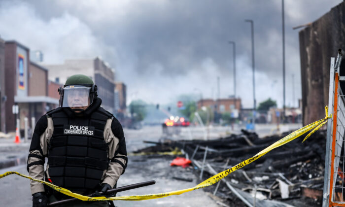 Un agente de policía permanece de pie entre el humo y los escombros mientras los edificios siguen ardiendo tras una noche de protestas y violencia después de la muerte de George Floyd, en Minneapolis, Minnesota, el 29 de mayo de 2020. (Charlotte Cuthbertson/The Epoch Times)