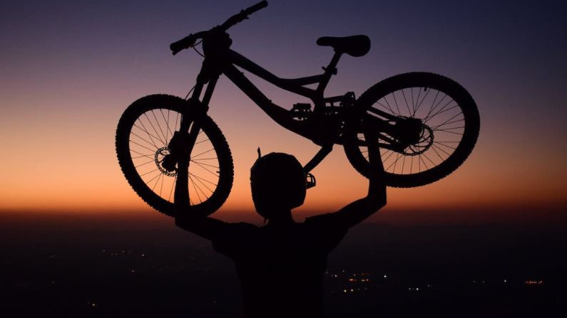 Concepto de éxito, logro, consecución y victoria con ciclista en bicicleta de montaña. Mujer feliz en bicicleta de montaña levantando los brazos alzando la bicicleta por el mar durante la puesta de sol animando y celebrando en la cima. (Pexels / Isabelli Pontes)