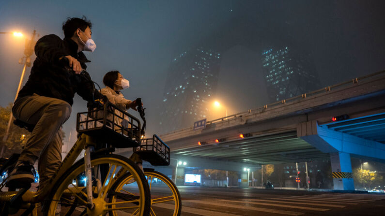 Una pareja lleva mascarillas protectoras para protegerse de la propagación del COVID-19 mientras se detienen en sus bicicletas compartidas para mirar hacia el edificio CCTV oscurecido por el smog y niebla el 20 de noviembre de 2022, en Beijing, China. (Foto de Kevin Frayer/Getty Images)
