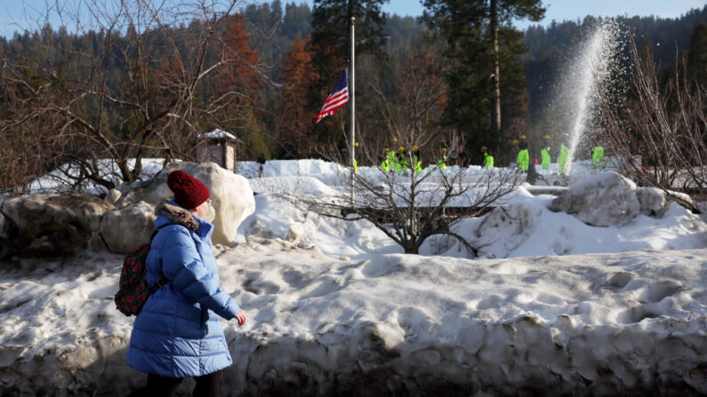 Una peatona camina cuando miembros de la Guardia Nacional del Ejército de California Fuerza de Tarea Conjunta Rattlesnake (ARRIBA R) palan nieve de un tejado después de una serie de tormentas de invierno cayó más de 100 pulgadas de nieve en las montañas de San Bernardino en el sur de California el 8 de marzo de 2023 en Crestline, California. (Mario Tama/Getty Images)