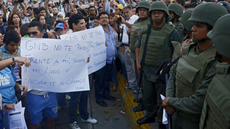  Manifestantes sostienen carteles que dicen "GNB no te pares frente a mí, párate a mi lado y sacude al cubano" frente a miembros de la Guardia Nacional en el bastión opositor de la plaza Altamira en Caracas el 17 de marzo de 2014. (LEO RAMIREZ/AFP via Getty Images)
