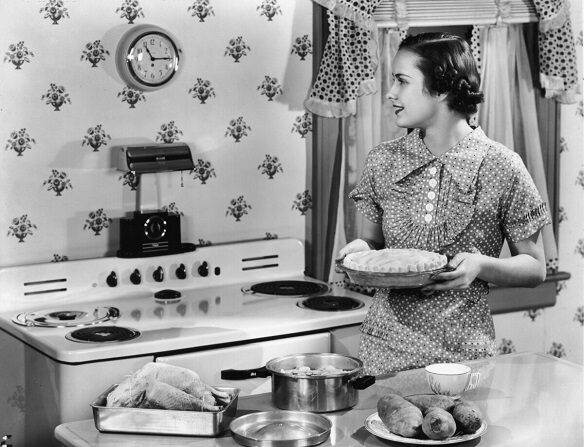 Un ama de casa sostiene un pastel sin hornear mientras mira un reloj de pared en una cocina. Sobre la encimera hay un pollo asado y patatas sin preparar. (Hulton Archive/Getty Images)
