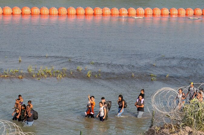 Migrantes caminan entre alambre de concertina y una serie de boyas colocadas en el agua a lo largo de la frontera del Río Grande con México, en Eagle Pass, Texas, el 16 de julio de 2023, para impedir la entrada de inmigración ilegal en Estados Unidos. (SUZANNE CORDEIRO/AFP vía Getty Images)