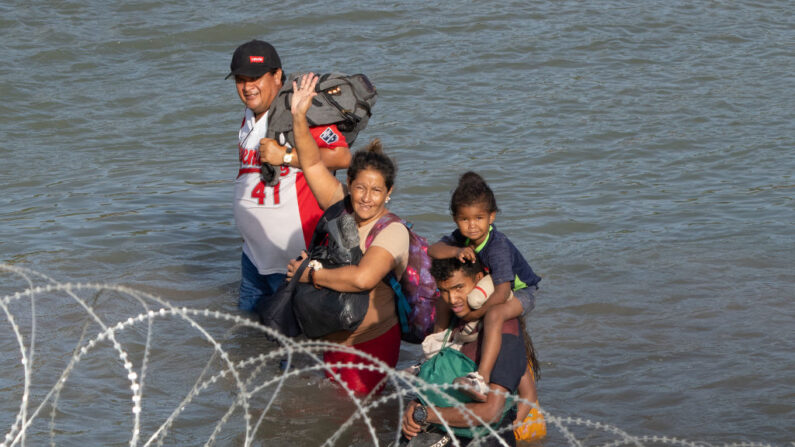 Migrantes saludan mientras caminan cerca de alambre de concertina en el agua a lo largo de la frontera del Río Grande con México en Eagle Pass, Texas, el 16 de julio de 2023. (SUZANNE CORDEIRO/AFP vía Getty Images)