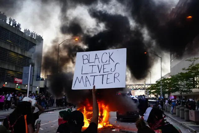 Un hombre sostiene un cartel de Black Lives Matter mientras un coche de policía arde frente a él durante una protesta frente al CNN Center en Atlanta, Georgia, el 29 de mayo de 2020, por la muerte en Minneapolis de George Floyd mientras estaba bajo custodia policial. (Elijah Nouvelage/Getty Images)