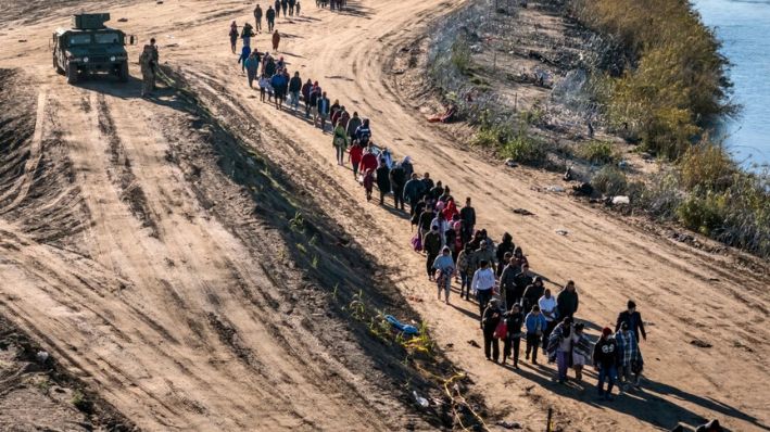 En esta vista aérea, un grupo de más de 1000 inmigrantes camina hacia un centro de procesamiento de campo de la Patrulla Fronteriza de EE. UU. después de cruzar el Río Grande desde México, el 18 de diciembre de 2023 en Eagle Pass, Texas. (John Moore/Getty Images)