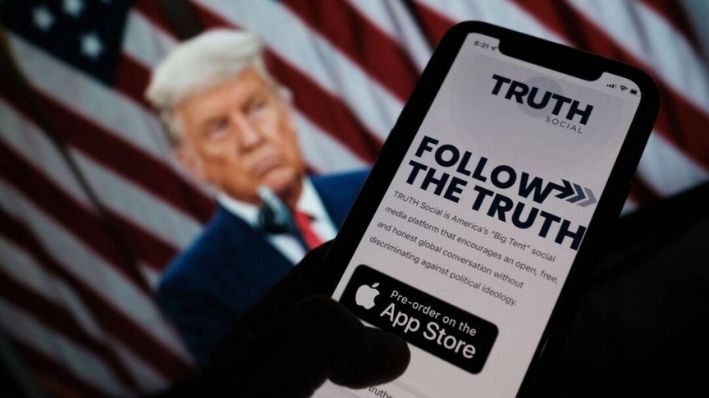 Una fotografía ilustrativa muestra a una persona revisando la tienda de aplicaciones de un teléfono inteligente buscando Truth Social con una foto del expresidente Donald Trump en la pantalla de una computadora al fondo, en Los Ángeles, el 20 de octubre de 2021. (CHRIS DELMAS/AFP a través de Getty Images)