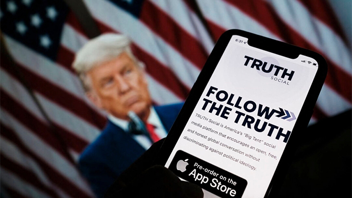 Esta foto ilustrativa muestra a una persona comprobando la tienda de aplicaciones de un smartphone en busca de Truth Social con una foto del expresidente Donald Trump en la pantalla de un ordenador al fondo, en Los Ángeles, el 20 de octubre de 2021. (Chris Delmas/AFP vía Getty Images)