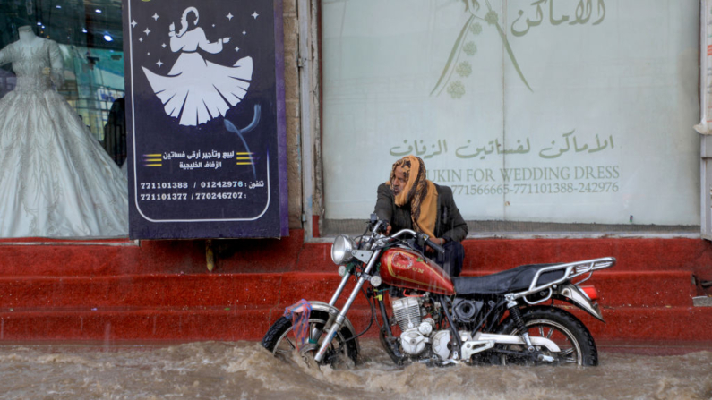 Un hombre sentado junto a su motocicleta en las escaleras de una tienda mientras el agua inunda una calle en medio de fuertes lluvias en Sanaa (Yemen), el 31 de marzo de 2023. (Mohammed Huwais/AFP vía Getty Images)