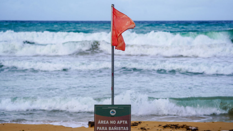 Una señal de advertencia del peligro bajo una bandera roja, indicando condiciones inseguras para nadar, se ven en la playa mientras las olas chocan contra la orilla en Luquillo, Puerto Rico, el 13 de agosto de 2024. (Jaydee Lee Serrano/AFP vía Getty Images)