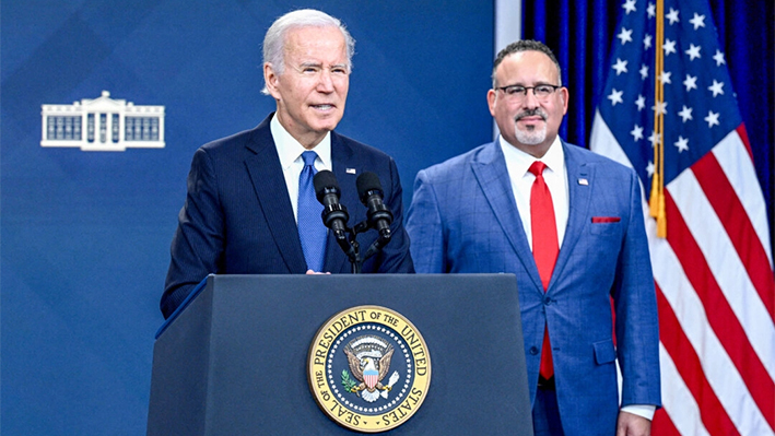 El secretario de Educación, Miguel Cardona (d), mira mientras el presidente Joe Biden pronuncia un discurso sobre la prueba beta del portal de alivio de la deuda estudiantil, en el Auditorio South Court del Edificio de Oficinas Ejecutivas Eisenhower, junto a la Casa Blanca, en Washington, el 17 de octubre de 2022. (Brendan Smialowski/AFP vía Getty Images)
