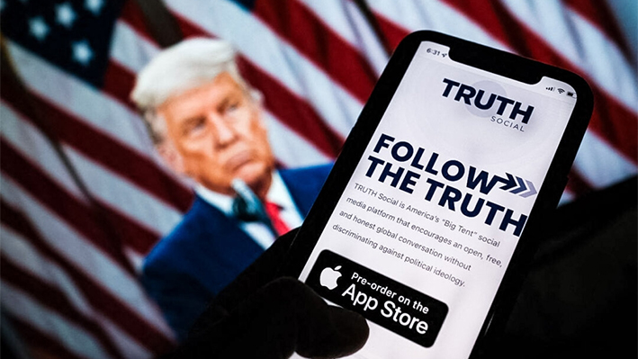 Esta foto ilustrativa muestra a una persona comprobando la tienda de aplicaciones de un smartphone en busca de Truth Social con una foto del expresidente Donald Trump en la pantalla de una computadora al fondo, en Los Ángeles, el 20 de octubre de 2021. (Chris Delmas/AFP vía Getty Images)
