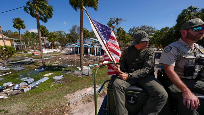 El oficial Nate Martir, agente de la ley de la Comisión de Pesca, Vida Silvestre y Conservación de Florida, sostiene una bandera estadounidense que yacía en el suelo entre escombros, mientras patrulla desde un buggy de pantano con capacidad para aguas altas, tras el paso del huracán Helene, en Cedar Key, Florida, el viernes 27 de septiembre de 2024. (AP Photo/Gerald Herbert)