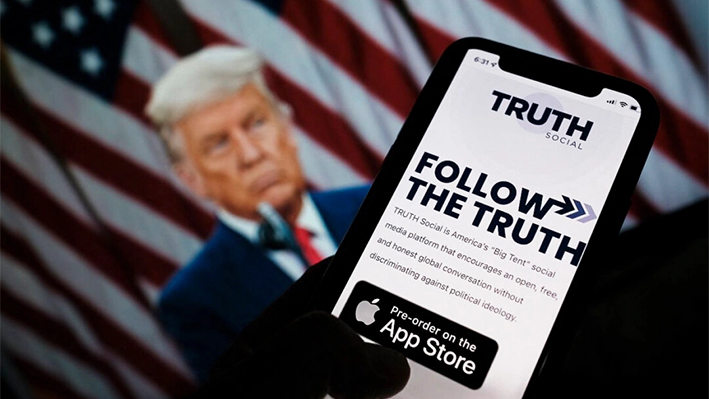 Foto ilustrativa de una persona comprobando la tienda de aplicaciones de un smartphone en busca de Truth Social con una foto del expresidente Donald Trump en la pantalla de un ordenador al fondo, en Los Ángeles el 20 de octubre de 2021. (Chris Delmas/AFP vía Getty Images)