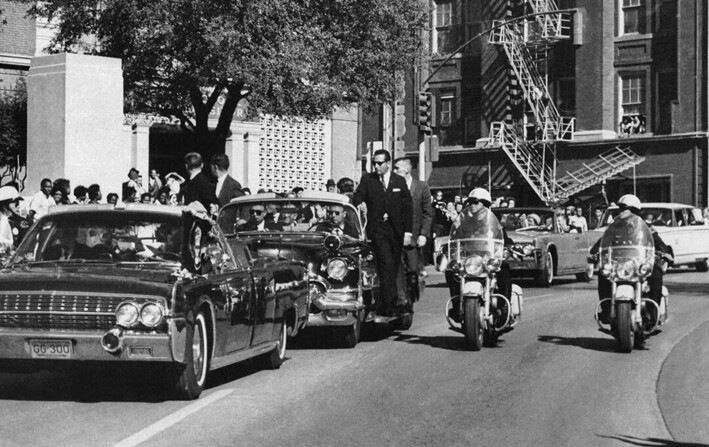 Visto a través del parabrisas del convertible en primer plano, la mano del presidente John F. Kennedy se extiende hacia su cabeza segundos después de recibir un disparo fatal mientras la primera dama Jacqueline Kennedy sostiene su antebrazo mientras la caravana avanza por Elm Street frente al Depósito de Libros Escolares de Texas, en Dallas, el 22 de noviembre de 1963. (James W. "Ike" Altgens/AP Photo)