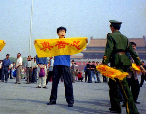 Um policial chinês se aproxima de um praticante do Falun Gong na Praça Tiananmen, em Pequim, enquanto ele segura uma faixa com os caracteres chineses de "verdade, compaixão e tolerância", princípios básicos do Falun Gong (via Minghui.org)